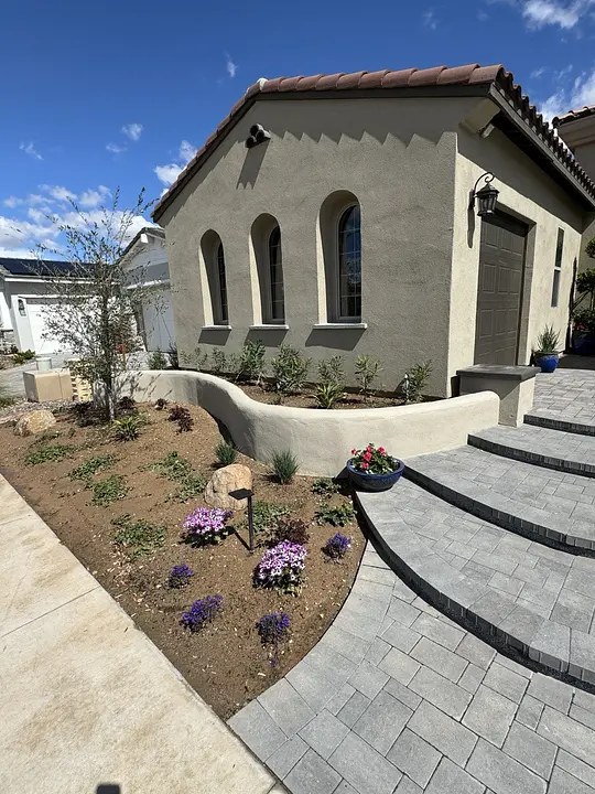 House with arched windows, gray pavers, and landscaped garden with purple and pink flowers under a clear sky.