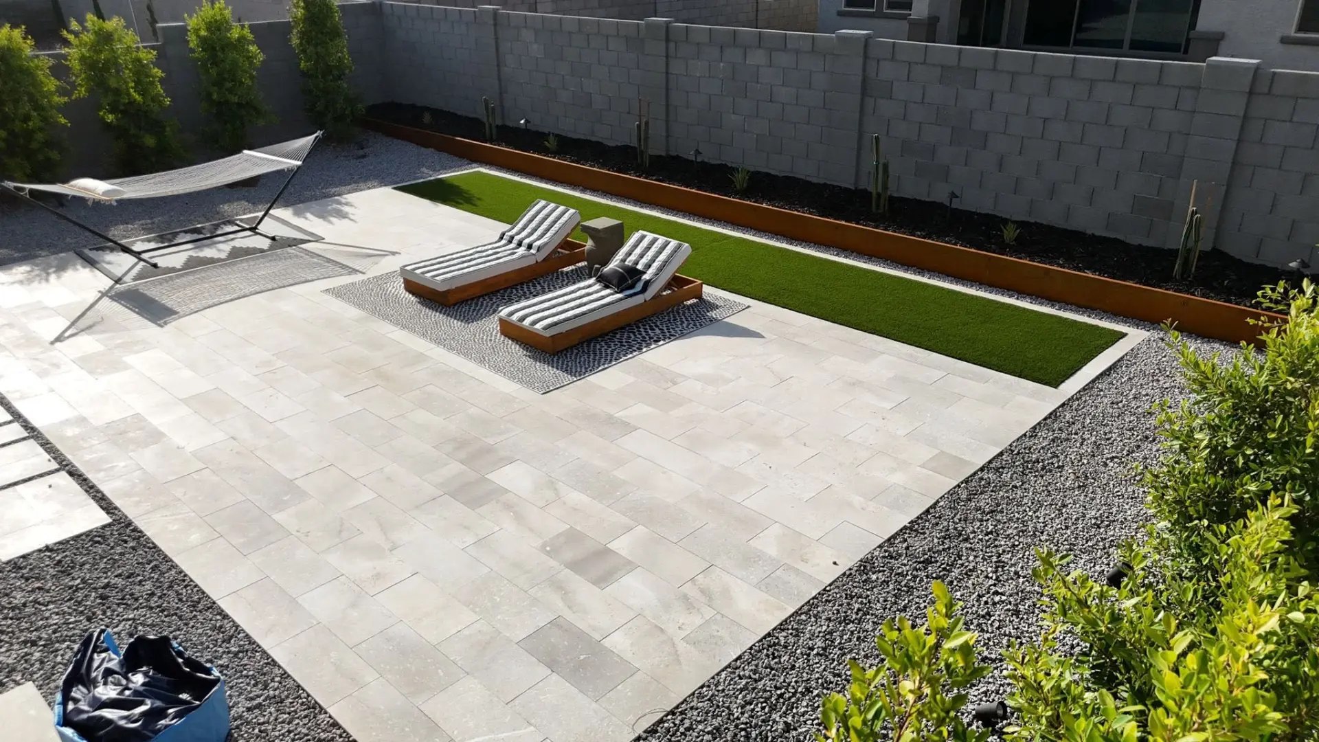 Small backyard with a light paver patio, a strip of artificial turf edged in concrete, gravel ground cover, a raised planting bed bordered by rusted steel edging, and two lounge chairs on an outdoor rug.