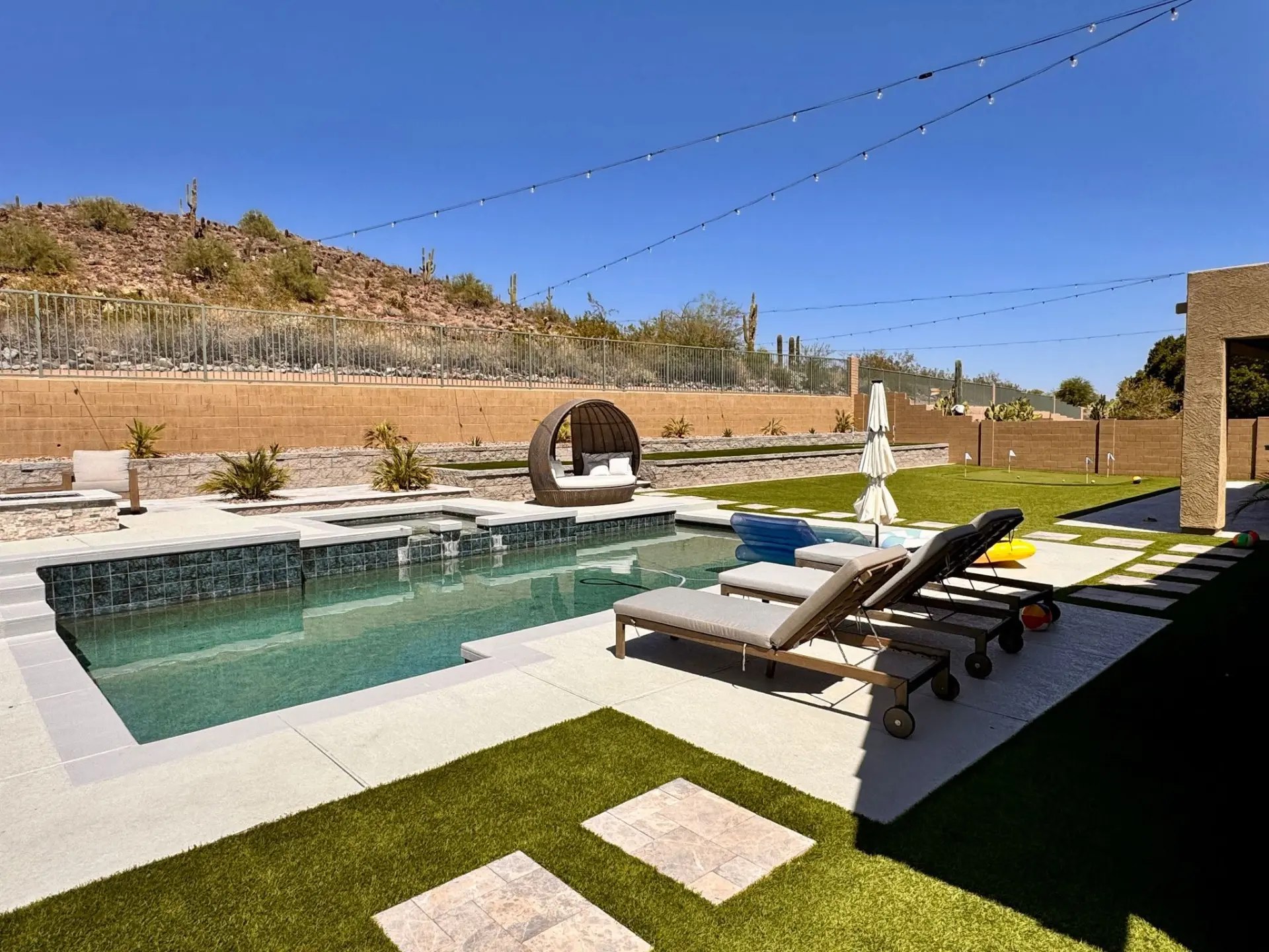Backyard pool with a raised spa spilling into the water, a light pool deck, turf laid with stepping pads, a putting green marked with flags, and two-tiered block retaining walls below a desert hillside.