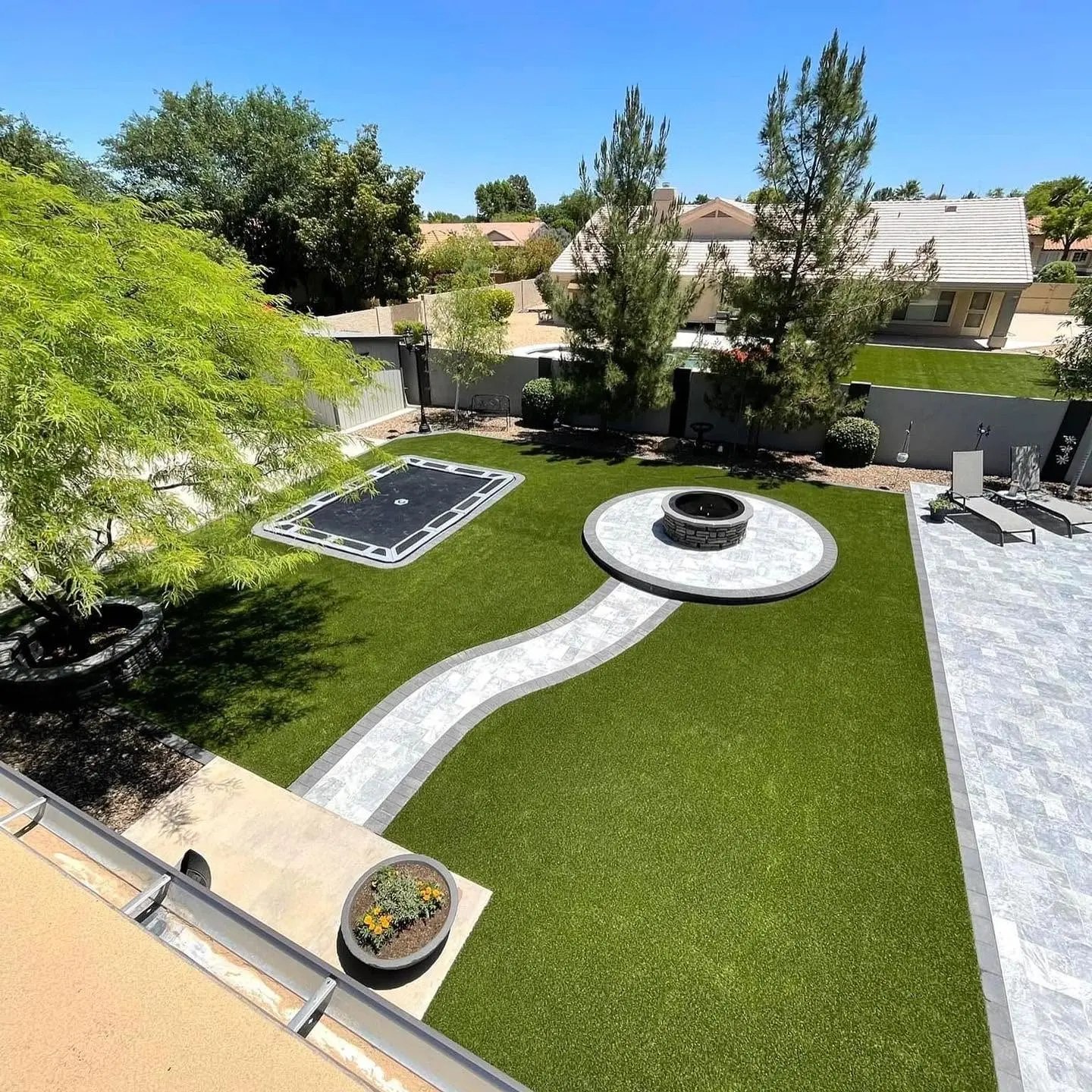 The same backyard seen from above: artificial turf with an in-ground trampoline, an S-curved paver walkway leading to a circular stacked-stone fire pit patio, a round stone tree ring, and a pool with a rock waterfall at the far wall.