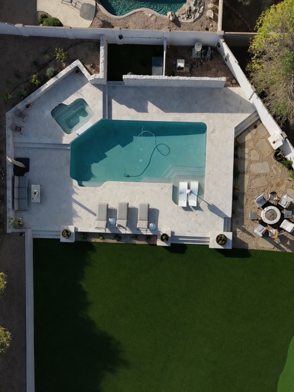 Aerial view of a backyard with a geometric pool holding a tanning shelf with two in-water loungers, a square raised spa in the upper corner, a light travertine paver deck in tiers with low capped retaining walls, a circular fire pit ringed by chairs on a flagstone pad, and artificial turf below.