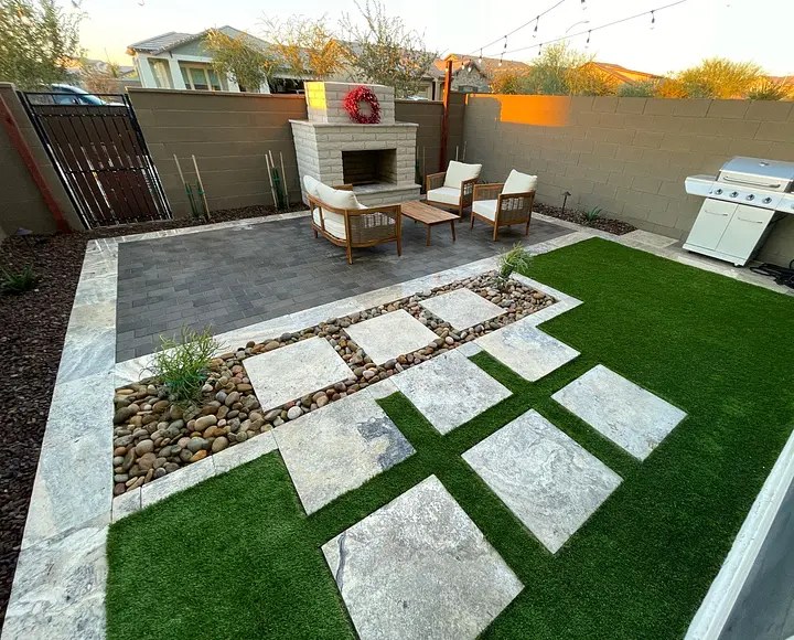 Modern patio with stone tiles, artificial grass, fireplace, chairs, and a grill, surrounded by a privacy wall.