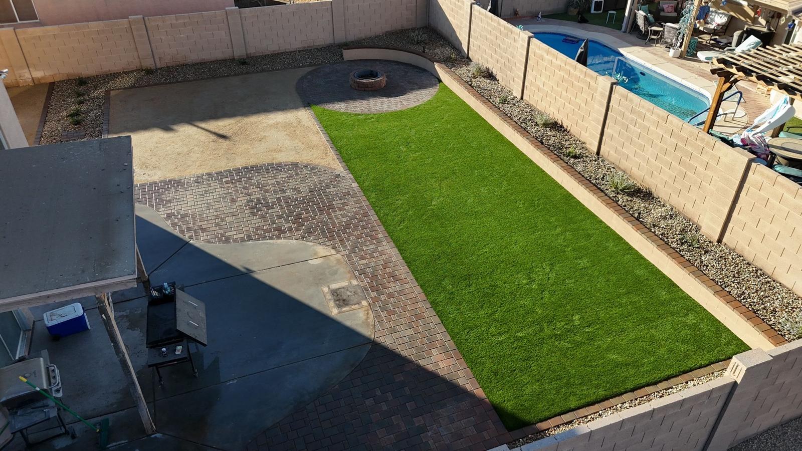 Finished backyard from above with a strip of artificial turf, multi-toned pavers around the concrete patio, a raised circular fire pit patio and a curved stone seat wall.