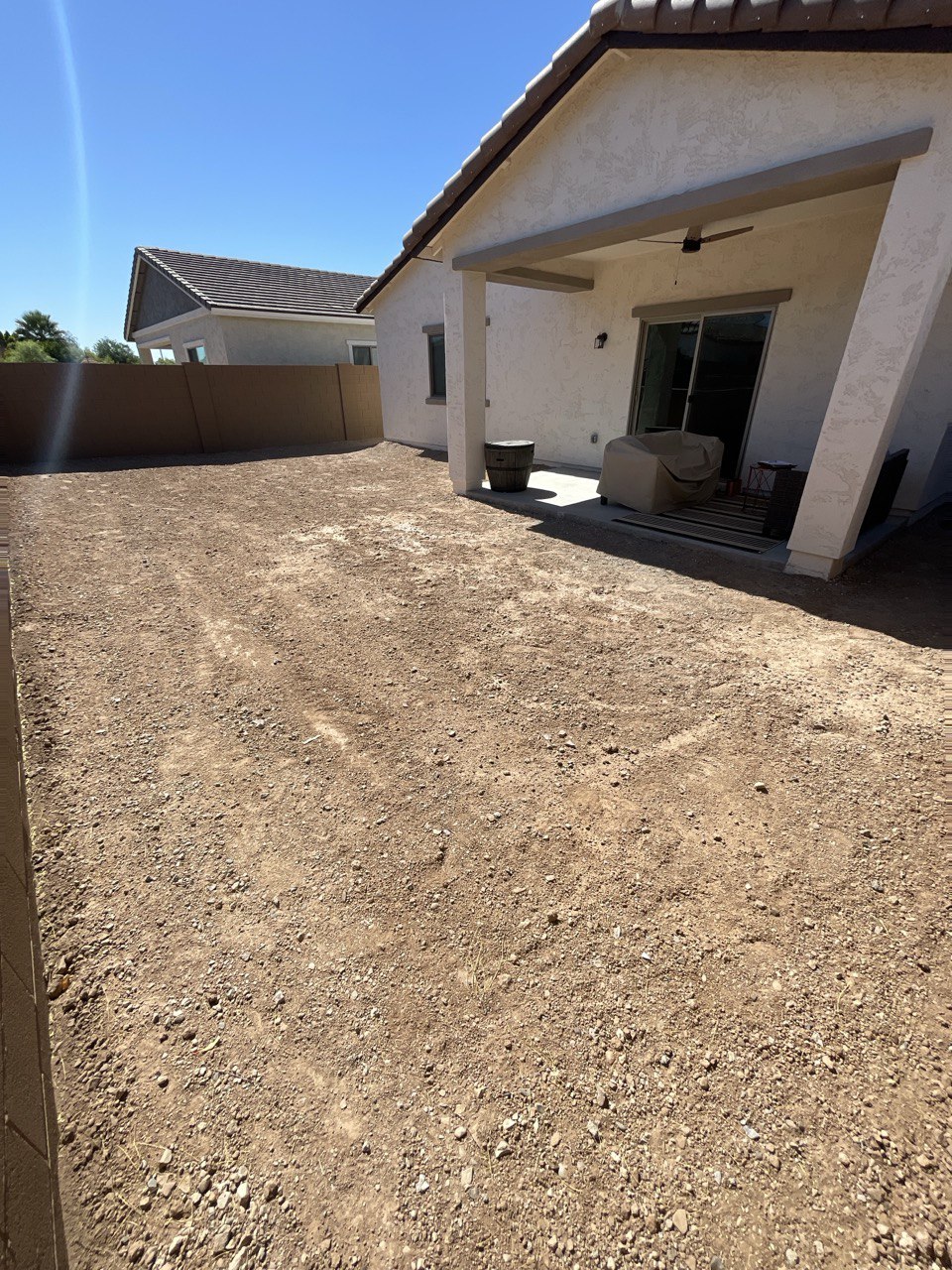 Backyard of bare dirt and gravel behind a stucco home with a covered patio, before hardscape work began.