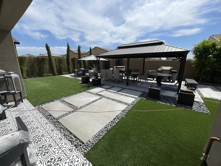 Modern backyard with patio, gazebo, seating, and grass. Stone paths and patterned tiles under a blue sky.