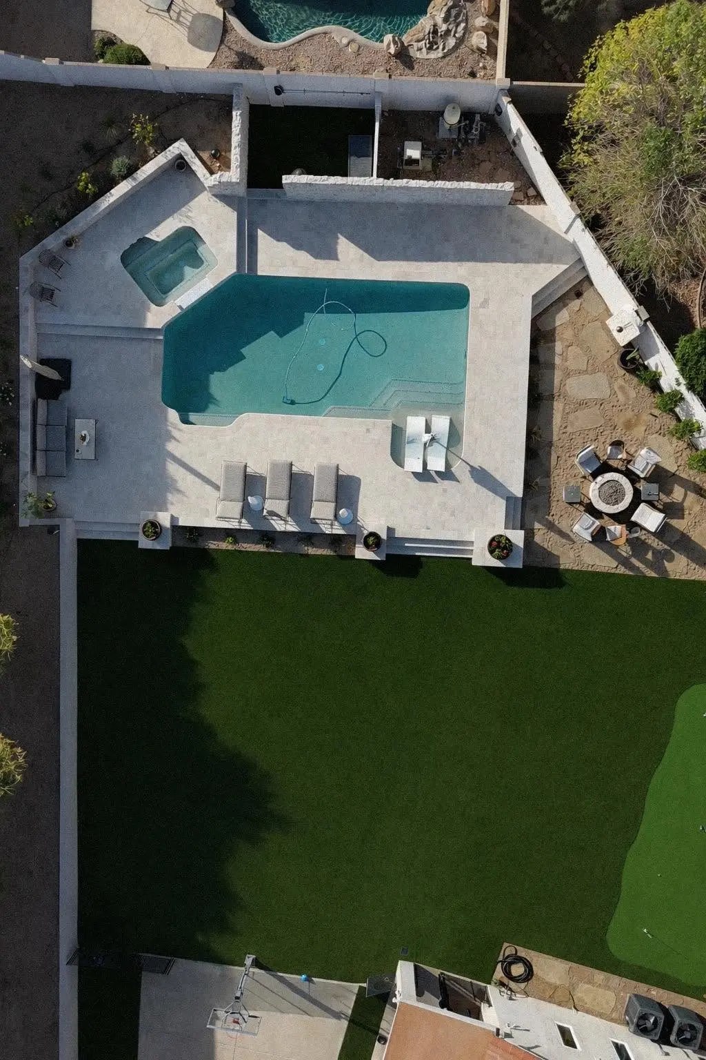 Aerial view of a backyard with a pool holding a tanning ledge and two in-water loungers, a light paver deck, artificial turf with a putting green, and a circular fire pit ringed by chairs on a flagstone pad.