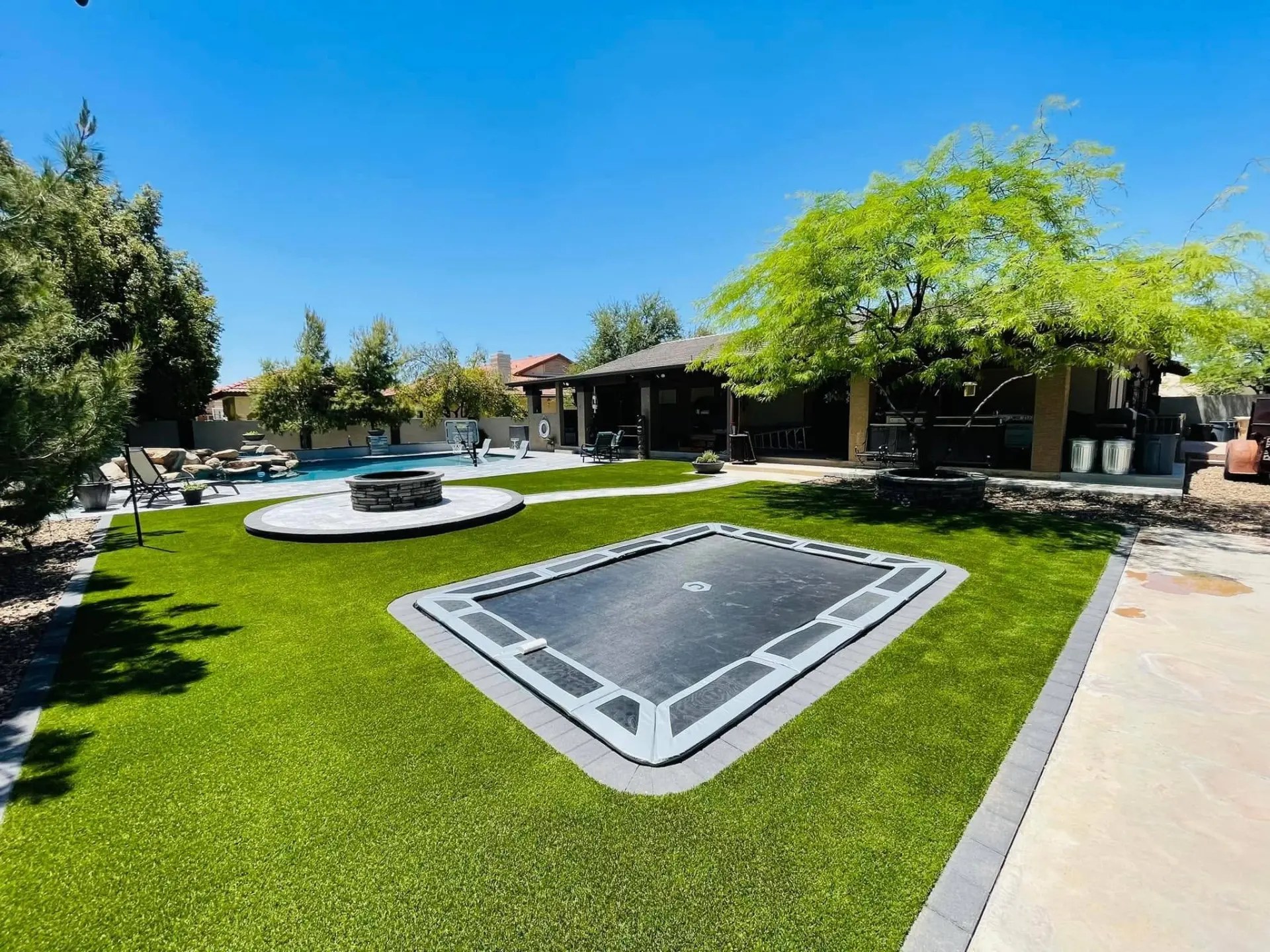 Backyard with an in-ground trampoline set into artificial turf, a raised circular fire pit patio with stacked stone edging, a stone tree ring beside a covered patio, and a pool with a rock waterfall in the background.