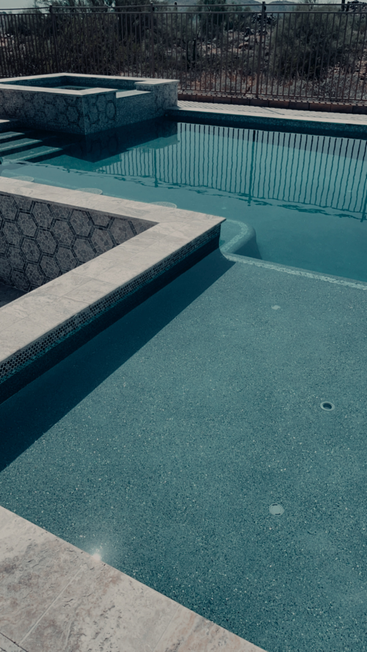New pool with a turquoise pebble finish, silver travertine coping, a raised spa wrapped in hexagon mosaic tile, and desert landscaping behind a wrought-iron fence.