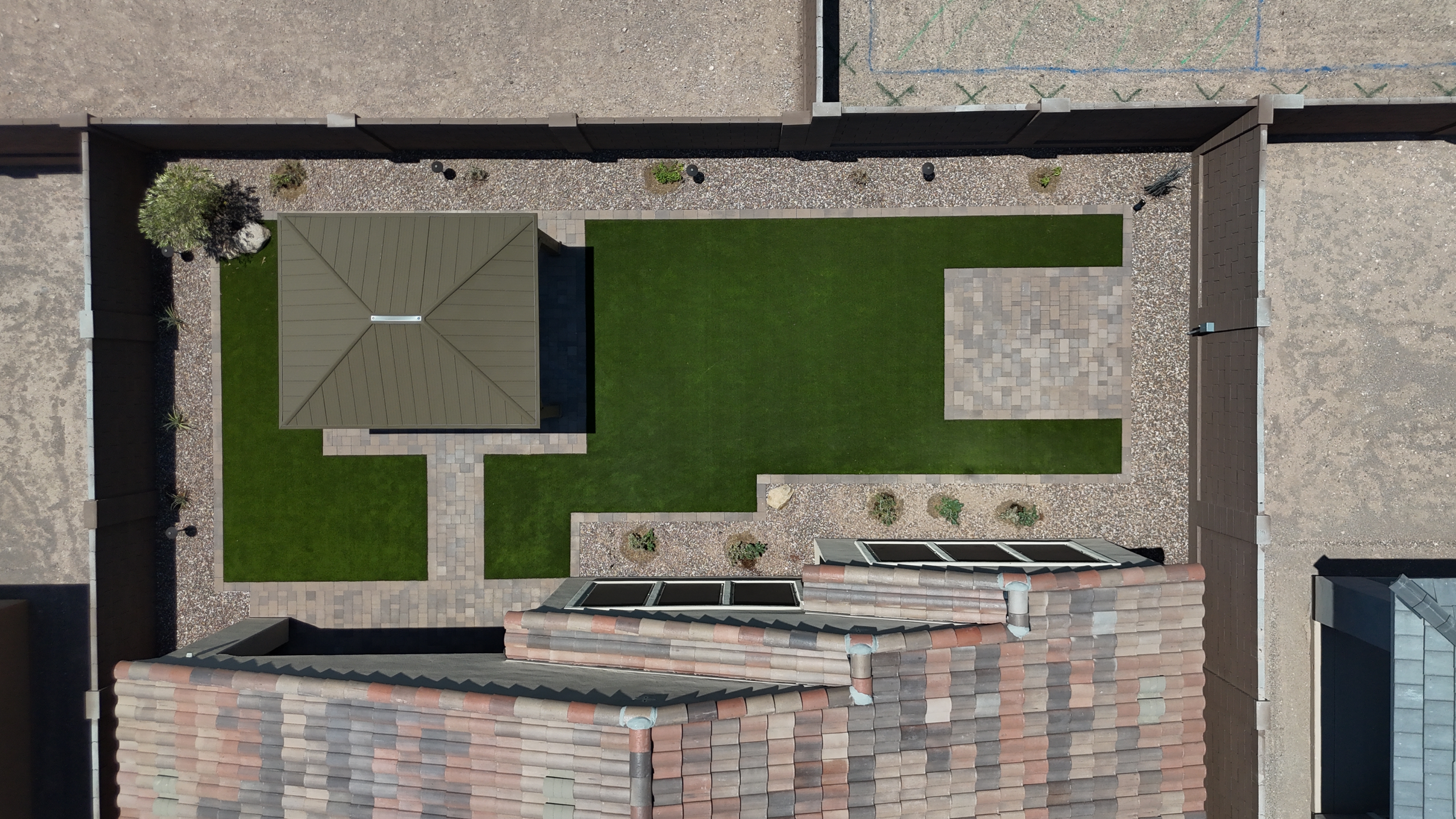 Finished backyard with artificial turf, a paver walkway to a dark roofed gazebo, a paver patio pad and a gravel planting bed with small shrubs and lights.