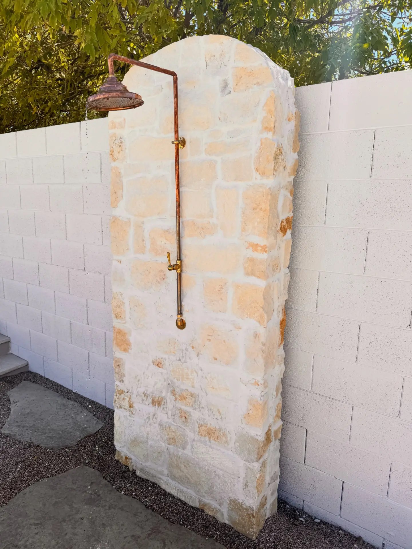 Outdoor shower built as a mortar-washed stone arch with copper pipe plumbing and a round rain head, standing over flagstone stepping stones set in dark gravel.