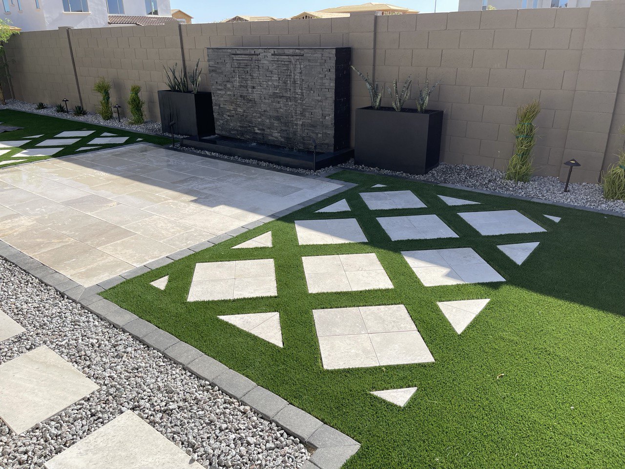Finished backyard with a light stone patio, artificial turf with a diamond pattern of stone pavers, a stacked stone water wall, black planters and landscape lights.