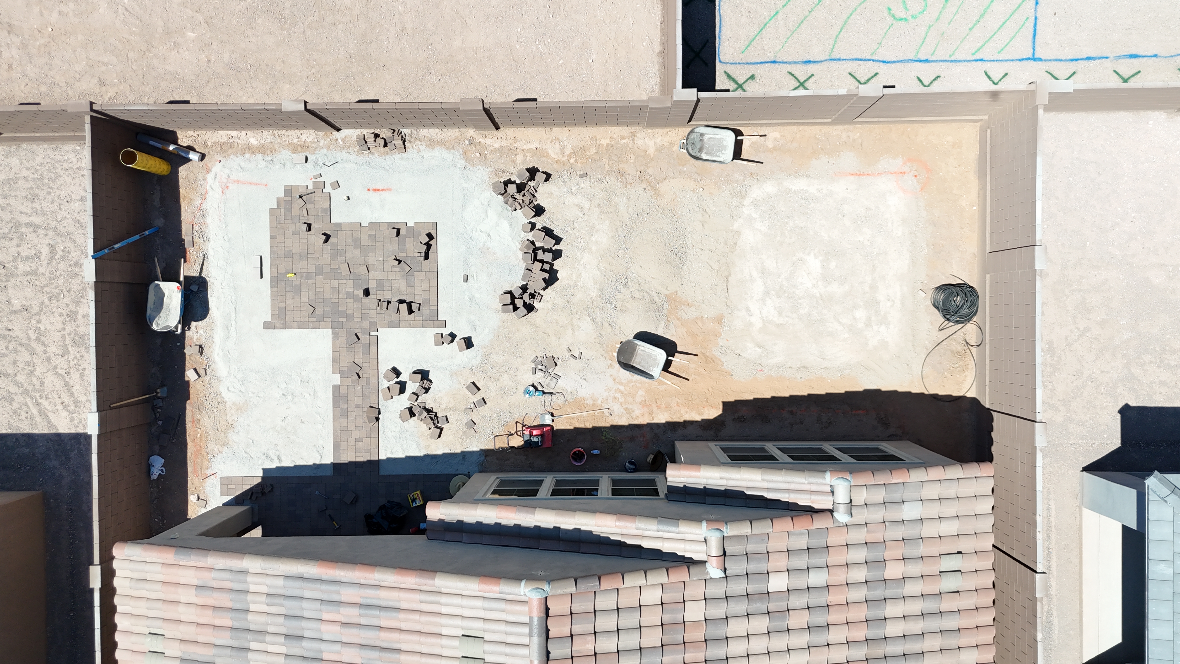 Backyard under construction with graded dirt, leveled sand bedding, partially laid pavers and wheelbarrows inside block walls.