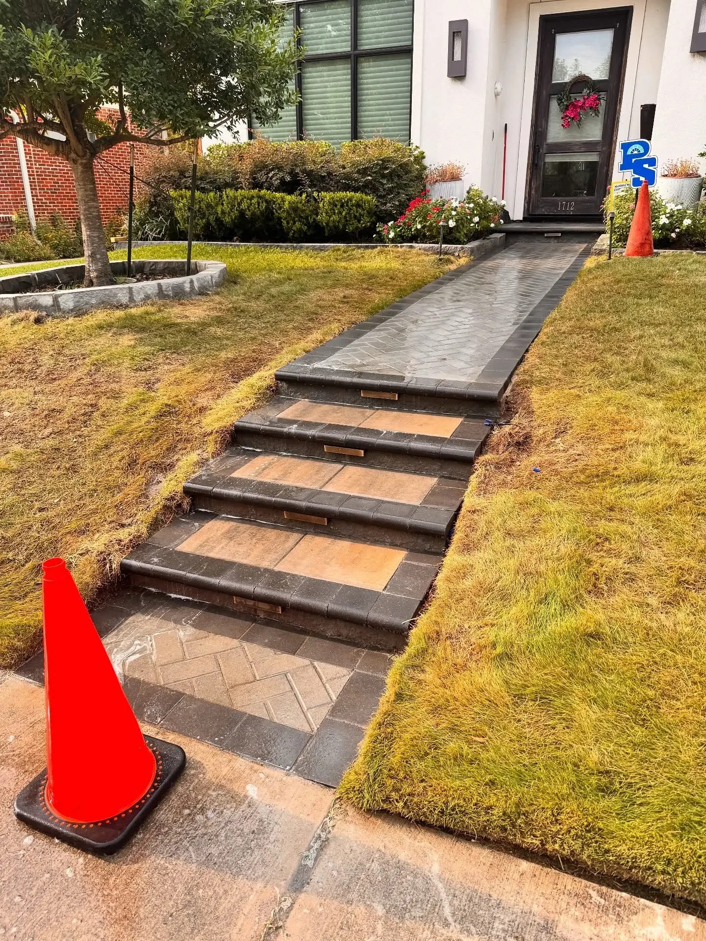 Rebuilt front steps and walkway with tan paver treads, dark charcoal coping, a herringbone paver path, brass step lights and a curved stone tree ring.