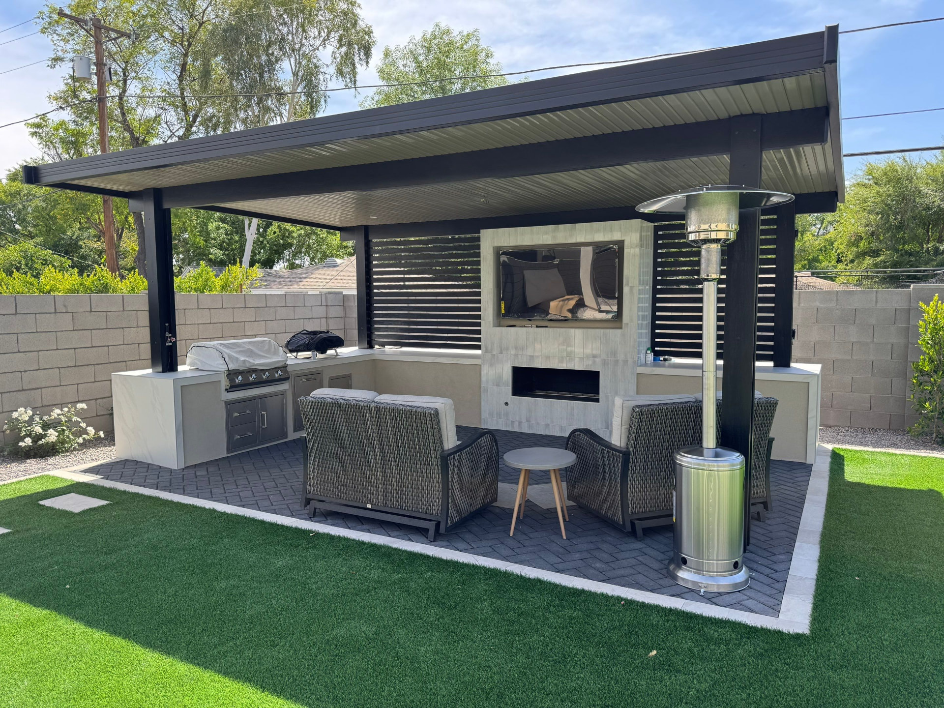 Backyard patio with a dark solid-roof pavilion, herringbone pavers edged in light gray, an outdoor kitchen with a built-in grill, and a stone feature wall holding a linear fireplace below a mounted TV, set in artificial turf.