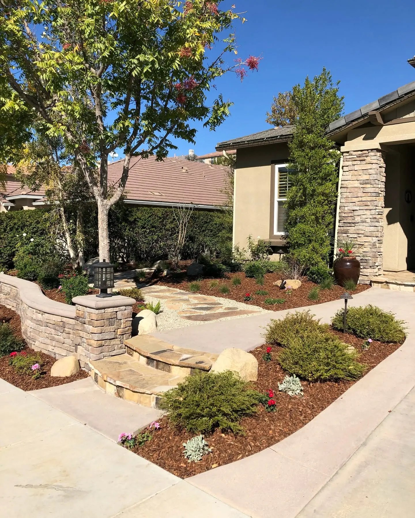 Front yard with a curved stacked-stone seat wall topped with smooth caps, a stone pillar holding a black lantern, natural stone steps, and a flagstone path running through gravel and mulched planting beds.