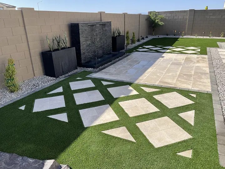 Modern backyard with geometric tile patterns on artificial grass, stone tiles, and a water feature against a tan wall.