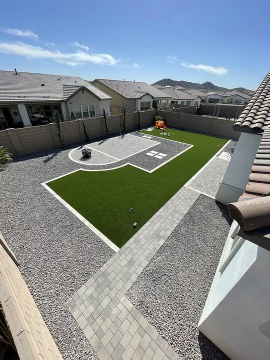 Modern backyard with artificial grass, paver patio, and play area, bordered by gravel and a low fence.