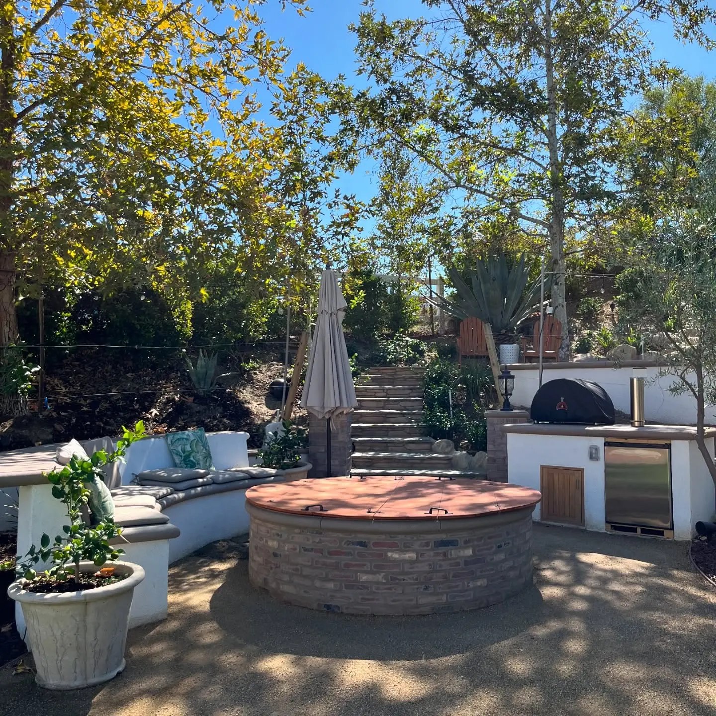 Hillside backyard with a round brick-faced fire pit under a wooden lid, a curved stucco bench with cushions, an outdoor kitchen island with an undercounter fridge and a covered pizza oven, and stone steps climbing to an upper patio.